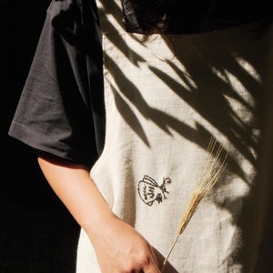May include: A beige apron featuring a small black embroidered graphic of a teapot on the front. The apron is worn over a black short-sleeved shirt and includes a front pocket. The fabric has a visible woven texture and a natural light color.