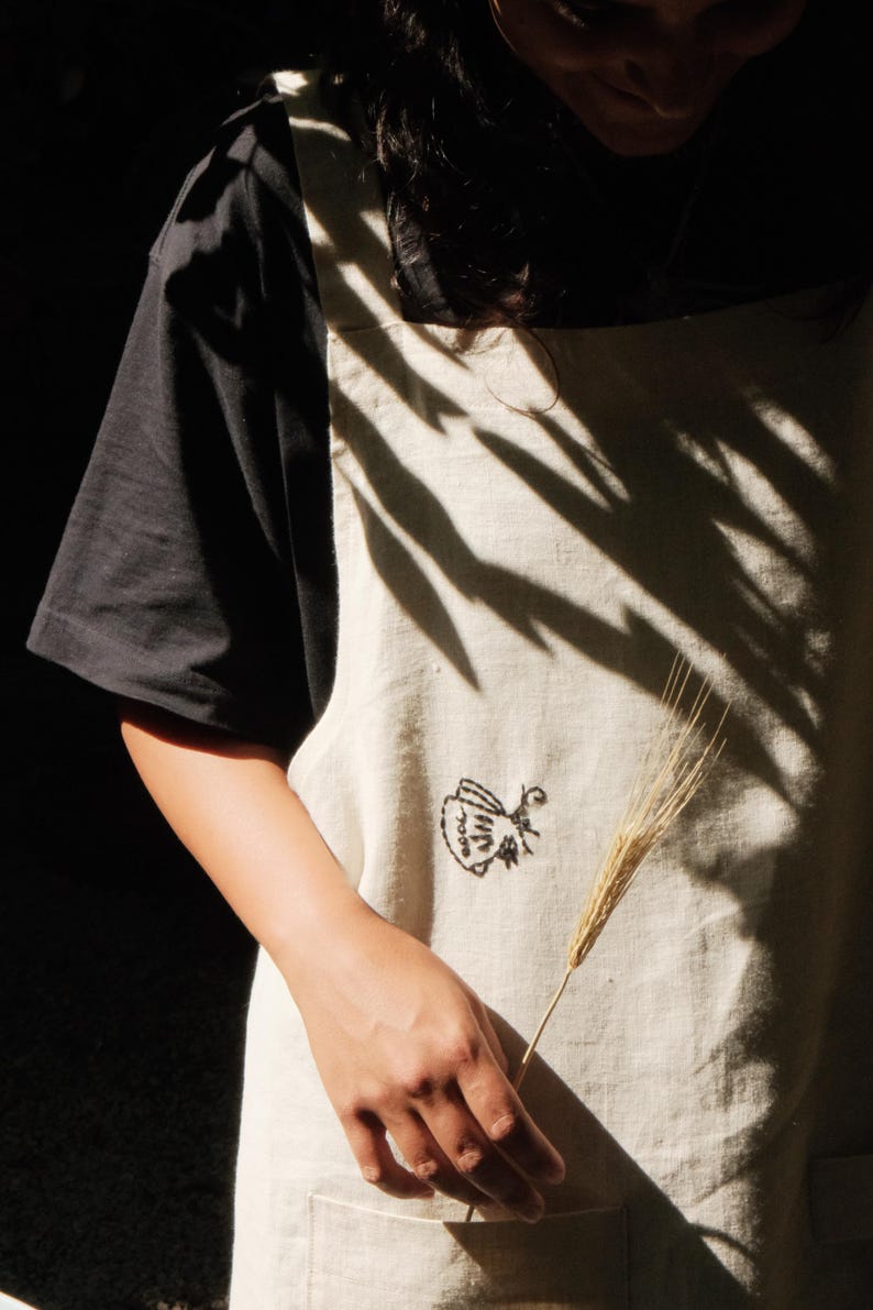 May include: A beige apron featuring a small black embroidered graphic of a teapot on the front. The apron is worn over a black short-sleeved shirt and includes a front pocket. The fabric has a visible woven texture and a natural light color.