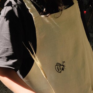 May include: A cream-colored apron featuring a small, black embroidered moth design on the front. The apron has wide shoulder straps and a visible pocket at the bottom, providing a simple and functional design for various tasks and activities.