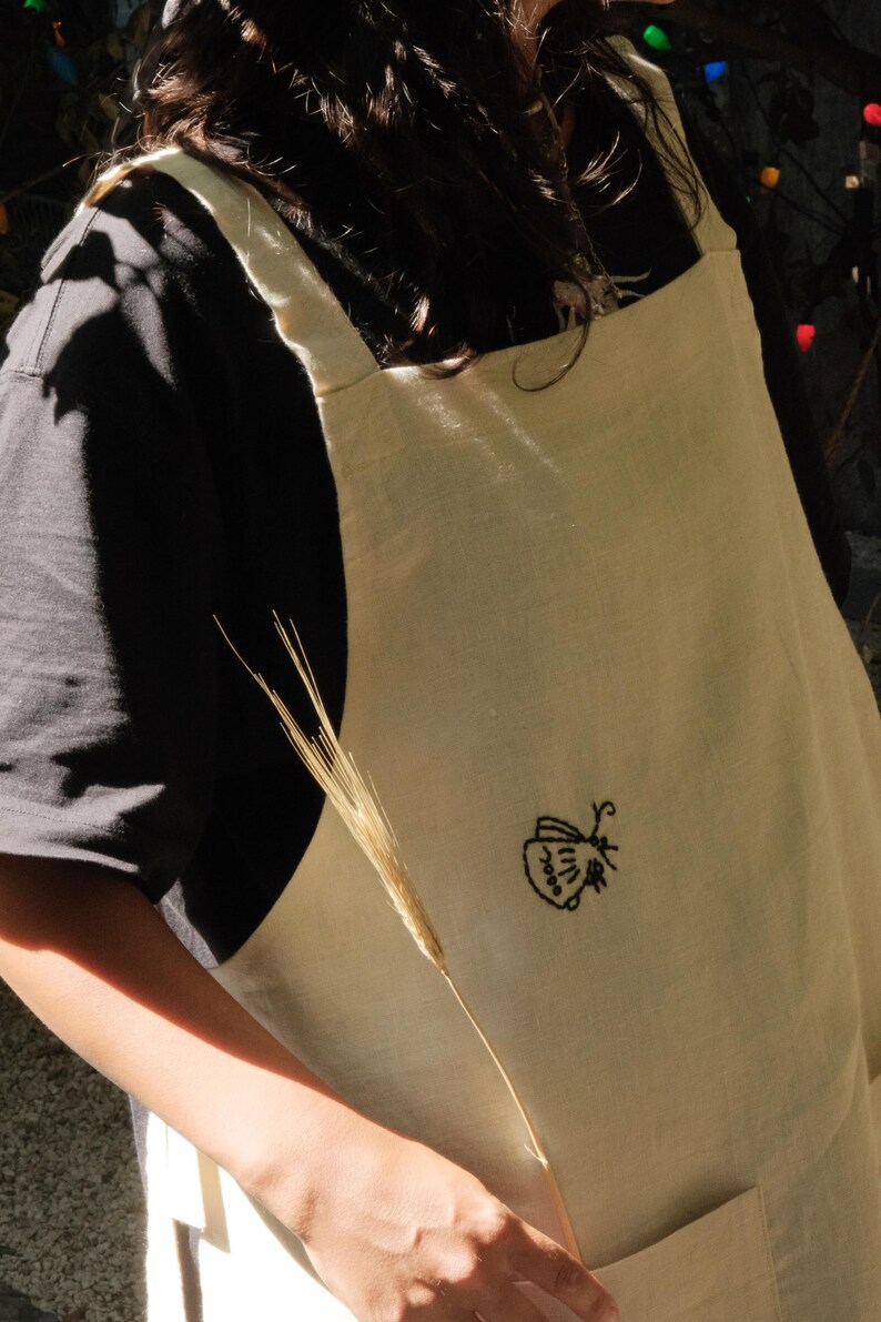 May include: A cream-colored apron featuring a small, black embroidered moth design on the front. The apron has wide shoulder straps and a visible pocket at the bottom, providing a simple and functional design for various tasks and activities.