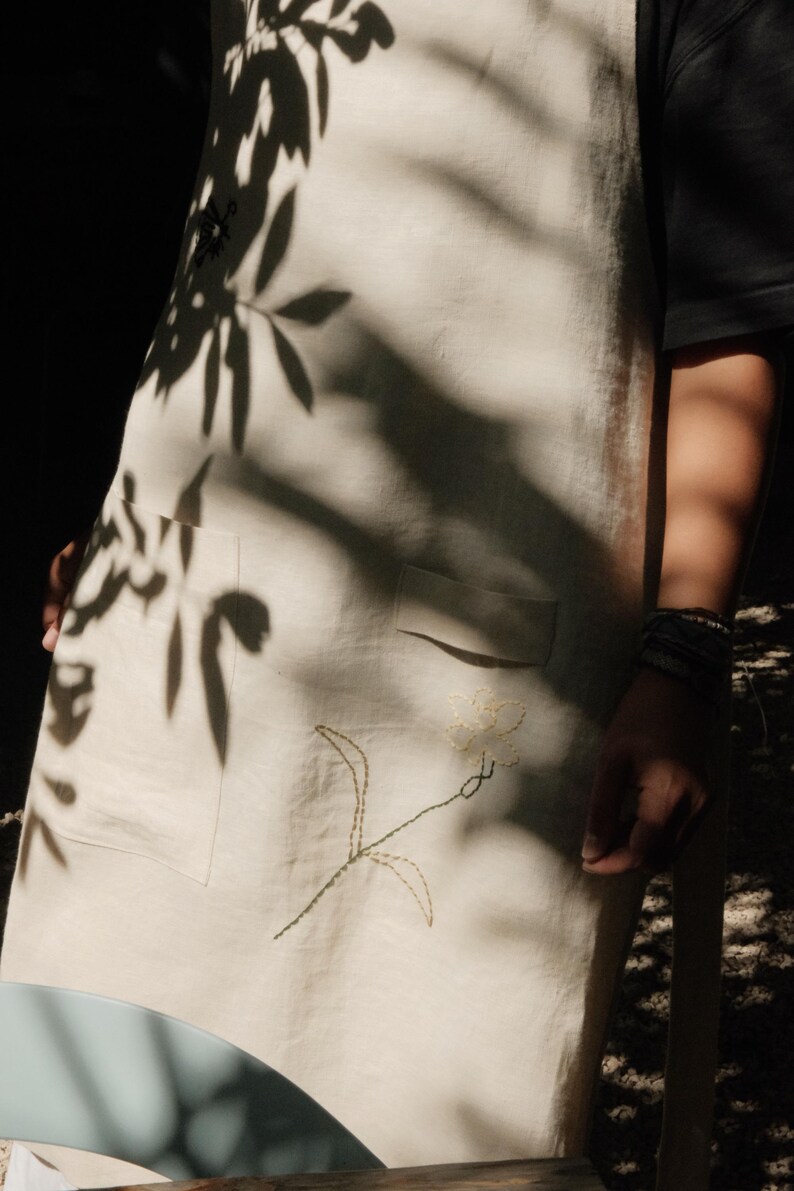 Op de afbeelding: Een cr&egrave;mekleurig schort met een delicaat, met de hand gestikt bloemenborduursel in lichtgroene en gele draad. Het schort is voorzien van een functionele voorzak en is gemaakt van een gladde, lichtgekleurde stof met een zichtbare weefstructuur.