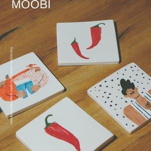 May include: Four square white ceramic tiles featuring hand-painted illustrations. Two tiles display single red chili peppers, one shows a person in an orange shirt holding blue jeans, and one depicts a person with dark hair against a black polka dot background. The text MŌOBI and FOR DESIGN LOVERS, WHO WANT SOMETHING DIFFERENT are visible.