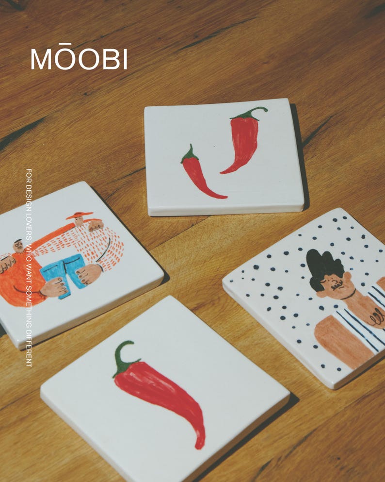 May include: Four square white ceramic tiles featuring hand-painted illustrations. Two tiles display single red chili peppers, one shows a person in an orange shirt holding blue jeans, and one depicts a person with dark hair against a black polka dot background. The text MŌOBI and FOR DESIGN LOVERS, WHO WANT SOMETHING DIFFERENT are visible.