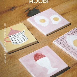 May include: Four square ceramic tiles featuring hand-painted designs: one yellow tile with a grid-patterned teapot, one pink tile with two fried eggs, one pink tile with a grid pattern and a fried egg, and one pink tile with a soft-boiled egg in a cup. Text reads MOOBI.