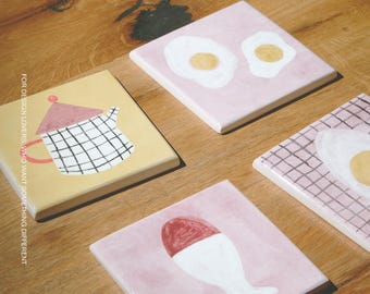Puede incluir: Cuatro azulejos de cerámica cuadrados con diseños pintados a mano: uno amarillo con una tetera de cuadrícula, uno rosa con dos huevos fritos, uno rosa con cuadrícula y un huevo frito, y uno rosa con un huevo pasado por agua. Texto: MOOBI.