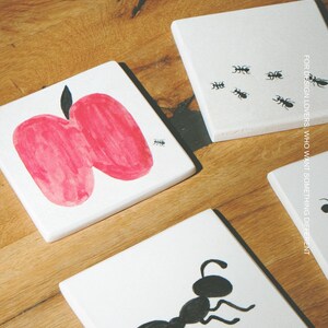 Pu&ograve; includere: Tre sottobicchieri quadrati in ceramica bianca con illustrazioni a inchiostro nero e rosso. Un sottobicchiere mostra una grande mela rossa con una minuscola formica nera, un altro una singola grande formica nera e il terzo presenta diverse piccole formiche nere.