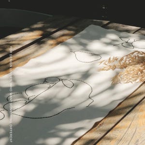 Pu&ograve; includere: Un runner da tavola in tessuto bianco con ricami a linee nere che raffigurano pere e forme astratte. Il testo MŌOBI e FOR DESIGN LOVERS, WHO WANT SOMETHING DIFFERENT &egrave; stampato sul tessuto. Un piccolo mazzetto di steli di grano essiccato &egrave; appoggiato sopra.