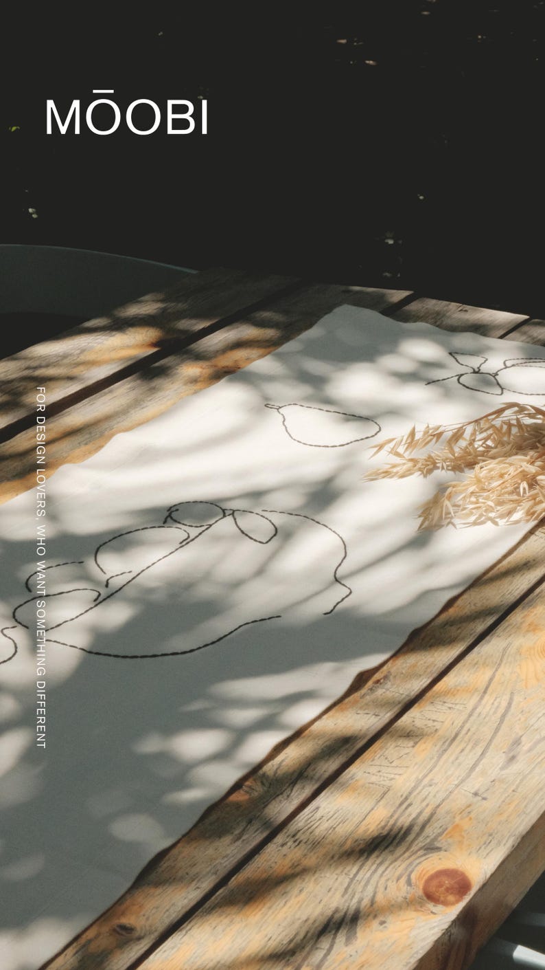 Pu&ograve; includere: Un runner da tavola in tessuto bianco con ricami a linee nere che raffigurano pere e forme astratte. Il testo MŌOBI e FOR DESIGN LOVERS, WHO WANT SOMETHING DIFFERENT &egrave; stampato sul tessuto. Un piccolo mazzetto di steli di grano essiccato &egrave; appoggiato sopra.