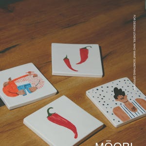 May include: Four square white ceramic coasters featuring colorful illustrations of red chili peppers and stylized figures. One coaster shows two red peppers, another a single pepper, and two depict illustrated characters. Text reads MŌOBI.