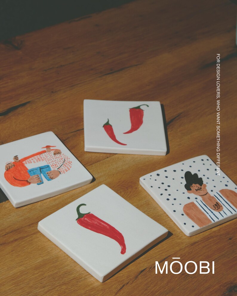 May include: Four square white ceramic coasters featuring colorful illustrations of red chili peppers and stylized figures. One coaster shows two red peppers, another a single pepper, and two depict illustrated characters. Text reads MŌOBI.