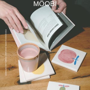 Puede incluir: Una taza de cer&aacute;mica rosa claro, una pila de posavasos cuadrados blancos con ilustraciones de frutas pintadas y un libro abierto con el texto MŌOBI y Clore Lounge Chair. El texto FOR DESIGN LOVERS, WHO WANT SOMETHING DIFFERENT es visible.