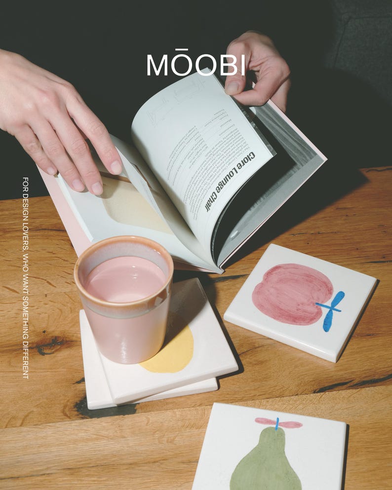 Puede incluir: Una taza de cer&aacute;mica rosa claro, una pila de posavasos cuadrados blancos con ilustraciones de frutas pintadas y un libro abierto con el texto MŌOBI y Clore Lounge Chair. El texto FOR DESIGN LOVERS, WHO WANT SOMETHING DIFFERENT es visible.