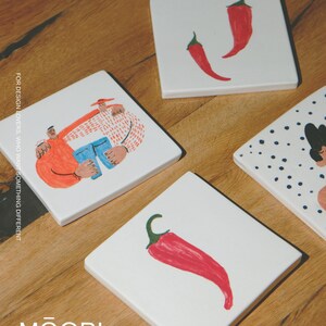 May include: Four square white ceramic coasters featuring colorful illustrations. One shows two figures embracing, another has two red chili peppers, one displays a single red chili pepper, and the fourth shows a partial figure with blue dots. Text reads MOOBI.