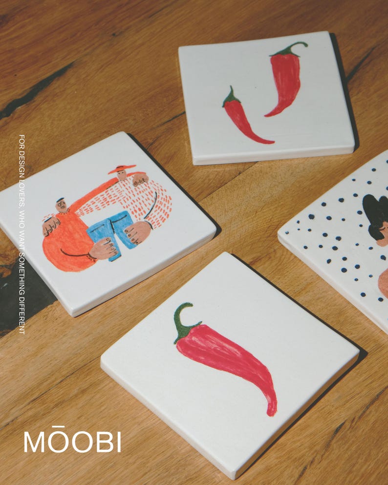 May include: Four square white ceramic coasters featuring colorful illustrations. One shows two figures embracing, another has two red chili peppers, one displays a single red chili pepper, and the fourth shows a partial figure with blue dots. Text reads MOOBI.