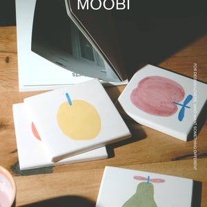 Puede incluir: Un juego de posavasos cuadrados de cer&aacute;mica blanca con ilustraciones de frutas pintadas a mano, que incluyen una manzana amarilla, una manzana roja y una pera verde, cada una con un tallo azul. El texto MŌOBI y FOR DESIGN LOVERS es visible.