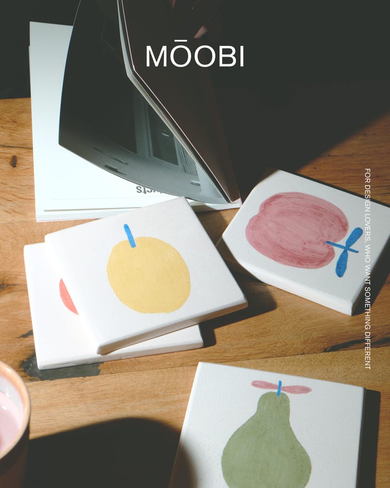 Puede incluir: Un juego de posavasos cuadrados de cer&aacute;mica blanca con ilustraciones de frutas pintadas a mano, que incluyen una manzana amarilla, una manzana roja y una pera verde, cada una con un tallo azul. El texto MŌOBI y FOR DESIGN LOVERS es visible.