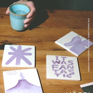 May include: Four square white ceramic coasters featuring hand-painted purple illustrations and text. Designs include a star shape, a volcano with text, a purple abstract form with text, and the words IT WAS FANTASTIC in purple lettering.