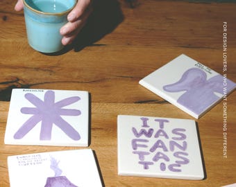 Puede incluir: Cuatro posavasos cuadrados de cerámica blanca con ilustraciones y texto pintados a mano en color morado. Los diseños incluyen una forma de estrella, un volcán con texto, una forma abstracta morada con texto y el texto IT WAS FANTASTIC.