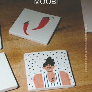 May include: Two square white ceramic tiles with hand-painted illustrations. One tile features two red chili peppers. The other tile depicts a person with dark hair wearing a blue and white striped shirt against a background of blue dots. Text reads MŌOBI.