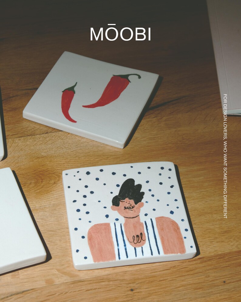 May include: Two square white ceramic tiles with hand-painted illustrations. One tile features two red chili peppers. The other tile depicts a person with dark hair wearing a blue and white striped shirt against a background of blue dots. Text reads MŌOBI.