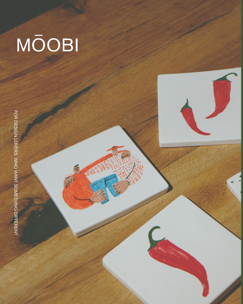 May include: Three square white ceramic coasters. One coaster features a colorful illustration of two figures embracing, while the other two coasters each display a single red chili pepper. The text MŌOBI and FOR DESIGN LOVERS, WHO WANT SOMETHING DIFFERENT is visible.