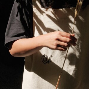 May include: An off-white apron featuring a small black embroidered graphic of a teapot. The apron has a square neckline, thick shoulder straps, and a visible front pocket. The fabric has a textured appearance and is worn over a black t-shirt.
