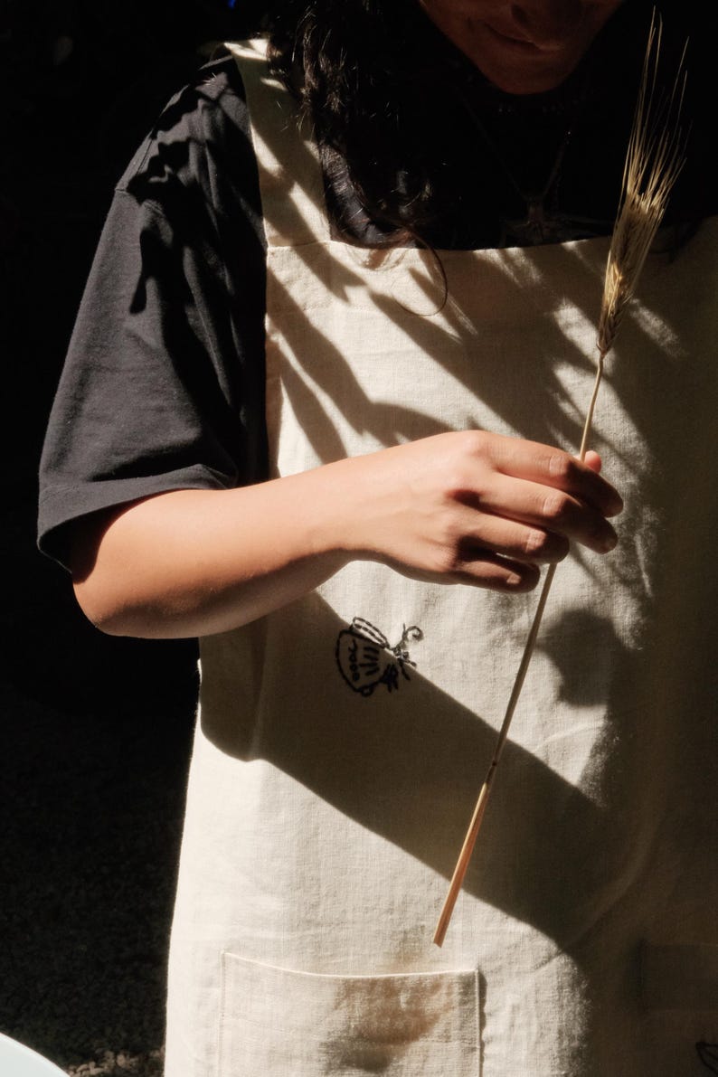 May include: An off-white apron featuring a small black embroidered graphic of a teapot. The apron has a square neckline, thick shoulder straps, and a visible front pocket. The fabric has a textured appearance and is worn over a black t-shirt.