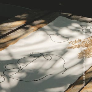 Pu&ograve; includere: Un runner da tavola rettangolare in tessuto bianco con ricami a linee nere che raffigurano una pera e un volto. Il testo MŌOBI &egrave; stampato in alto, mentre il testo FOR DESIGN LOVERS WHO WANT SOMETHING DIFFERENT &egrave; stampato verticalmente sul lato destro.