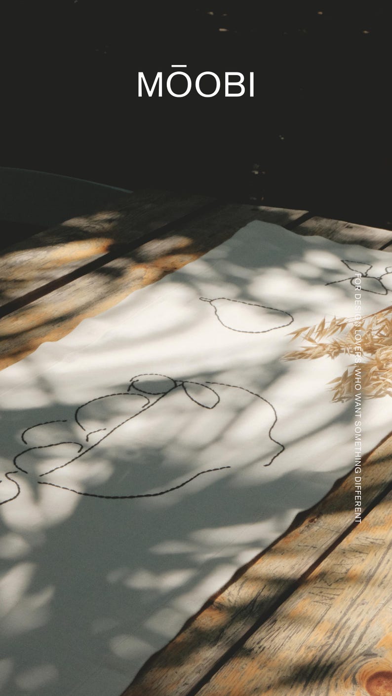 Pu&ograve; includere: Un runner da tavola rettangolare in tessuto bianco con ricami a linee nere che raffigurano una pera e un volto. Il testo MŌOBI &egrave; stampato in alto, mentre il testo FOR DESIGN LOVERS WHO WANT SOMETHING DIFFERENT &egrave; stampato verticalmente sul lato destro.