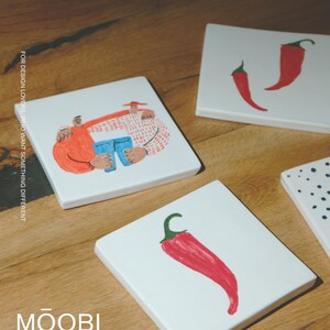 May include: Three square white ceramic coasters featuring hand-painted illustrations: one shows two figures embracing while holding blue jeans, another displays two red chili peppers, and the third features a single red chili pepper. Text reads MOOBI.