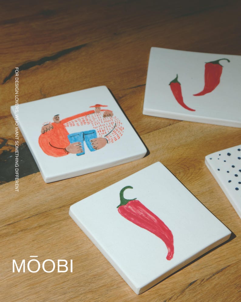 May include: Three square white ceramic coasters featuring hand-painted illustrations: one shows two figures embracing while holding blue jeans, another displays two red chili peppers, and the third features a single red chili pepper. Text reads MOOBI.