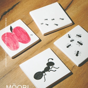 May include: Four square white coasters featuring black and white illustrations. One coaster displays a red apple, two feature small black ants, and one shows a single large black ant. The text "FOR DESIGN LOVERS, WHO WANT SOMETHING DIFFERENT" and "MŌOBI" are visible.