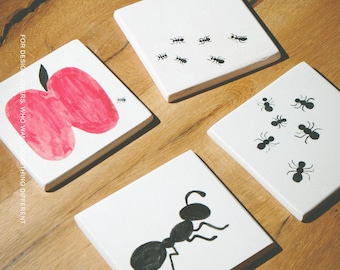 Puede incluir: Cuatro posavasos cuadrados blancos con ilustraciones en blanco y negro. Un posavasos muestra una manzana roja, dos muestran pequeñas hormigas negras y uno muestra una sola hormiga negra grande. El texto "FOR DESIGN LOVERS, WHO WANT SOMETHING DIFFERENT" y "MŌOBI" son visibles.