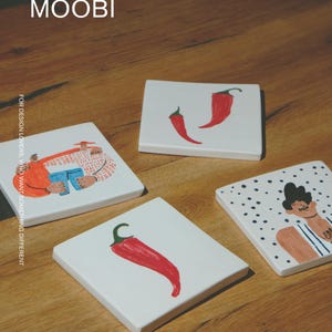 May include: Four square white ceramic coasters featuring hand-painted illustrations. Designs include two red chili peppers, a single red chili pepper, a person with dark hair on a polka dot background, and two figures embracing. Text reads MŌOBI.