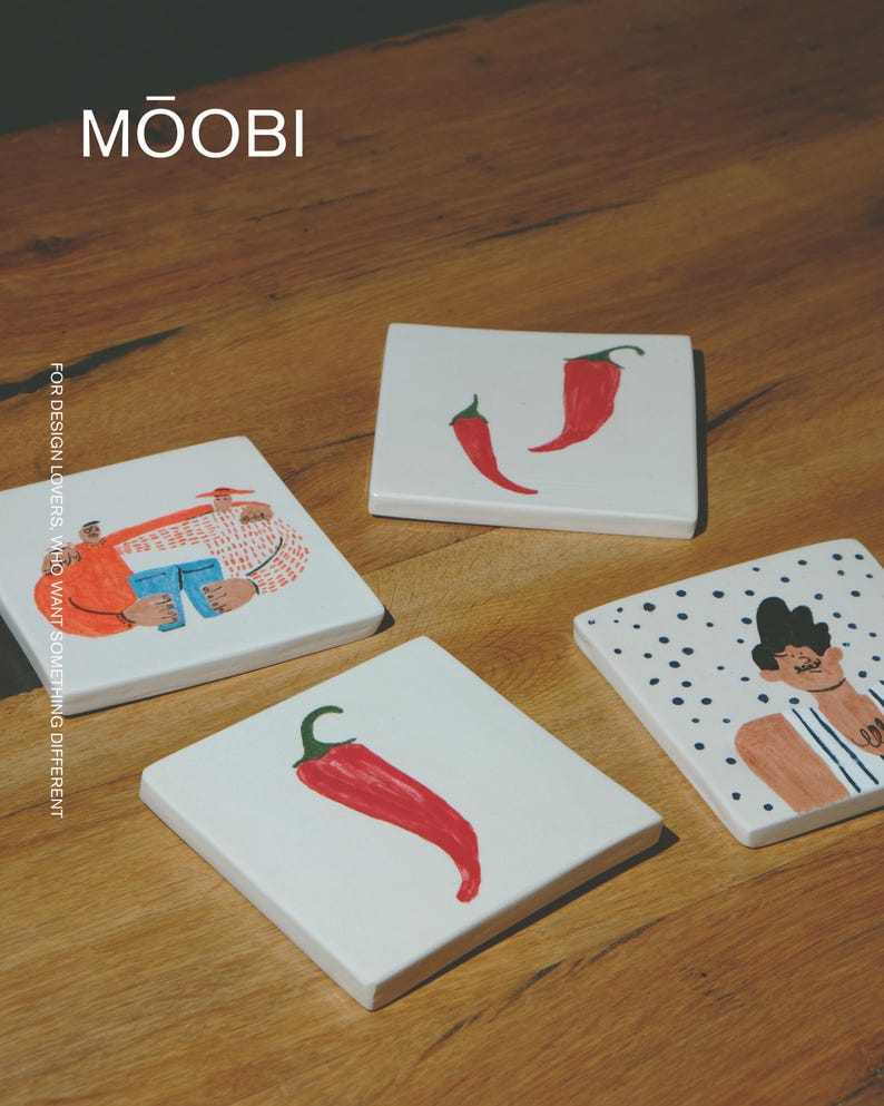 May include: Four square white ceramic coasters featuring hand-painted illustrations. Designs include two red chili peppers, a single red chili pepper, a person with dark hair on a polka dot background, and two figures embracing. Text reads MŌOBI.