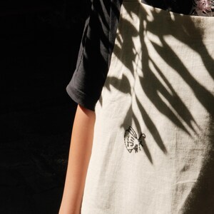 May include: An off-white apron featuring a small, black embroidered moth design on the chest. The apron has a simple, straight-cut bib with shoulder straps and a visible pocket at the bottom, worn over a plain black short-sleeved shirt.