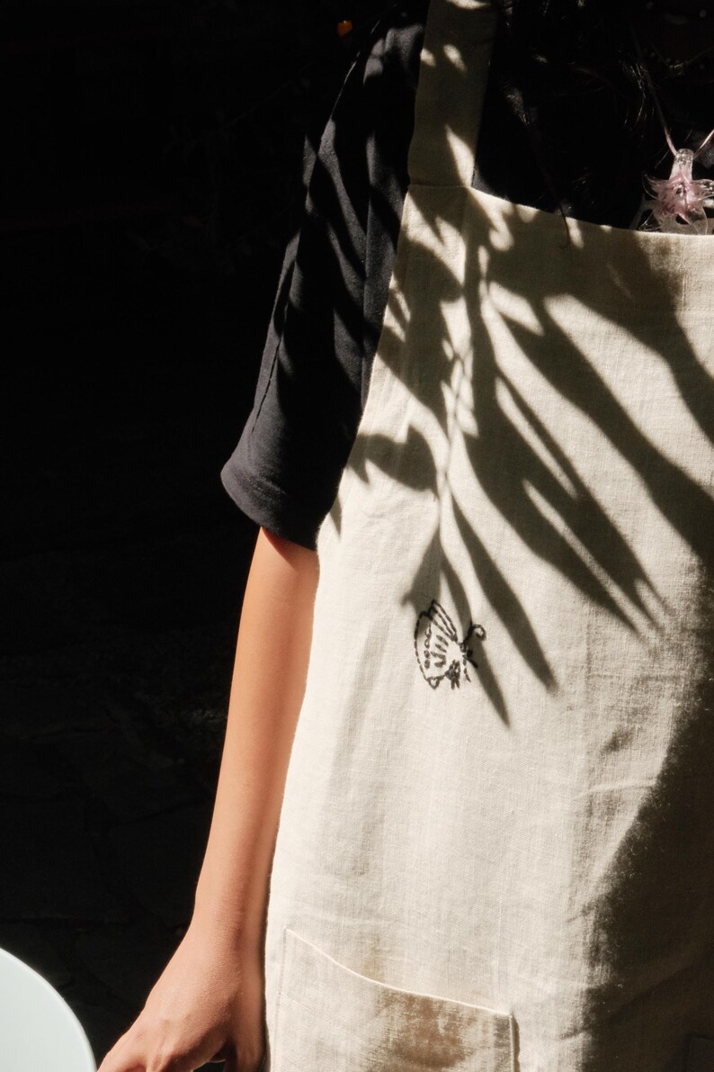 May include: An off-white apron featuring a small, black embroidered moth design on the chest. The apron has a simple, straight-cut bib with shoulder straps and a visible pocket at the bottom, worn over a plain black short-sleeved shirt.