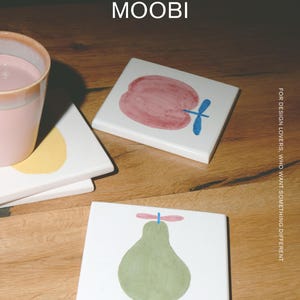 Puede incluir: Posavasos cuadrados de cer&aacute;mica blanca con ilustraciones de frutas pintadas a mano. Un posavasos muestra una manzana roja con un tallo azul, otro una pera verde con un tallo rosa. El texto MŌOBI y FOR DESIGN LOVERS, WHO WANT SOMETHING DIFFERENT es visible.