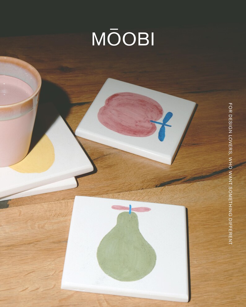 Puede incluir: Posavasos cuadrados de cer&aacute;mica blanca con ilustraciones de frutas pintadas a mano. Un posavasos muestra una manzana roja con un tallo azul, otro una pera verde con un tallo rosa. El texto MŌOBI y FOR DESIGN LOVERS, WHO WANT SOMETHING DIFFERENT es visible.