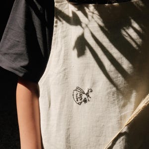 May include: An off-white apron featuring a small, black embroidered butterfly design on the front. The apron has wide shoulder straps and a large front pocket, layered over a plain black short-sleeved shirt, with natural light casting shadows.