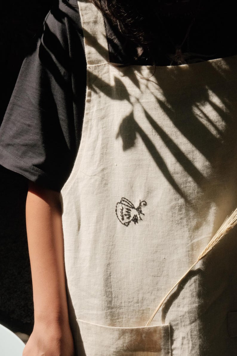 May include: An off-white apron featuring a small, black embroidered butterfly design on the front. The apron has wide shoulder straps and a large front pocket, layered over a plain black short-sleeved shirt, with natural light casting shadows.