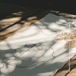 Pu&ograve; includere: Un runner da tavola rettangolare in tessuto bianco con ricami a linee nere che raffigurano una pera e un volto. Il testo MŌOBI &egrave; stampato in alto, mentre il testo FOR DESIGN LOVERS WHO WANT SOMETHING DIFFERENT &egrave; stampato verticalmente sul lato destro.