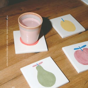 Puede incluir: Una taza de cer&aacute;mica rosa claro descansa sobre un posavasos cuadrado blanco con una ilustraci&oacute;n de una pera verde. Hay otros dos posavasos cuadrados blancos cerca, uno con una fruta amarilla y otro con una manzana rosa. El texto dice FOR DESIGN LOVERS, WHO WANT SOMETHING DIFFERENT y MŌOBI.