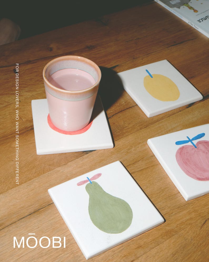 Puede incluir: Una taza de cer&aacute;mica rosa claro descansa sobre un posavasos cuadrado blanco con una ilustraci&oacute;n de una pera verde. Hay otros dos posavasos cuadrados blancos cerca, uno con una fruta amarilla y otro con una manzana rosa. El texto dice FOR DESIGN LOVERS, WHO WANT SOMETHING DIFFERENT y MŌOBI.