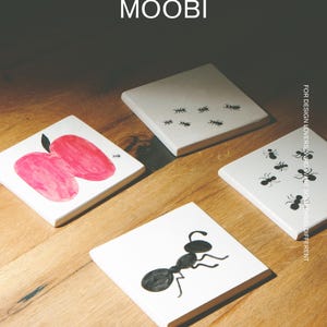 Pu&ograve; includere: Quattro sottobicchieri quadrati bianchi con grafiche nere e rosse. Uno presenta una mela rossa, uno una grande formica nera e due presentano motivi di piccole formiche nere. Il testo MŌOBI e FOR DESIGN LOVERS, WHO WANT SOMETHING DIFFERENT &egrave; visibile.
