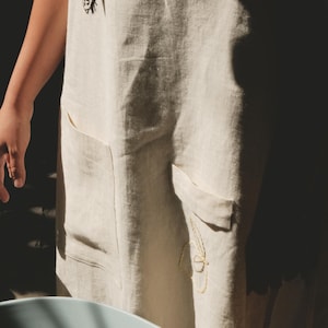 May include: A beige apron featuring two front pockets and embroidered details. The upper chest area displays a small black embroidered bee, while the lower section near the right pocket features a delicate yellow embroidered floral outline.