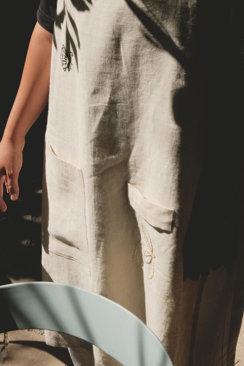 May include: A beige apron featuring two front pockets and embroidered details. The upper chest area displays a small black embroidered bee, while the lower section near the right pocket features a delicate yellow embroidered floral outline.