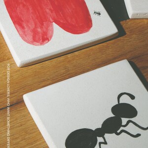 Pu&ograve; includere: Due sottobicchieri quadrati bianchi con illustrazioni dipinte a mano. Un sottobicchiere mostra una grande mela rossa con un picciolo nero e una piccola formica nera. L'altro mostra una grande formica nera. Il testo MOOBI &egrave; stampato in basso.