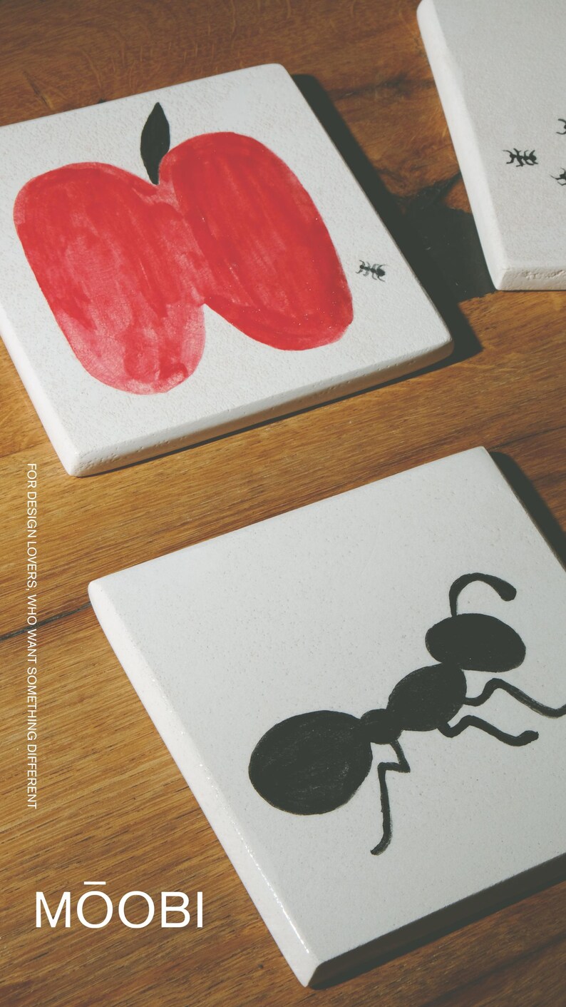 Pu&ograve; includere: Due sottobicchieri quadrati bianchi con illustrazioni dipinte a mano. Un sottobicchiere mostra una grande mela rossa con un picciolo nero e una piccola formica nera. L'altro mostra una grande formica nera. Il testo MOOBI &egrave; stampato in basso.