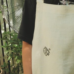 May include: An off-white apron featuring a small, black embroidered butterfly design on the front. The apron has wide shoulder straps and is worn over a black short-sleeved shirt. The fabric has a visible weave texture throughout the garment.